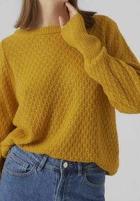 Senfgelber Strickpullover mit strukturiertem Muster, weiter Halslinie und verlängerten Ärmeln. Kombiniert mit hoch taillierter blauer Jeans.