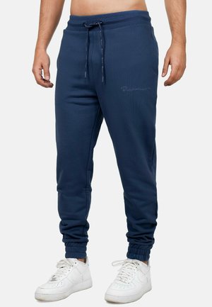 Uomo che indossa pantaloni da jogging blu navy con polsini elasticizzati e sneakers bianche, in piedi davanti a uno sfondo chiaro uniforme.