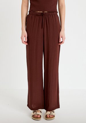 Donna che indossa pantaloni marroni a vita alta e taglio ampio con coulisse e sandali beige con fibbia, in piedi davanti a uno sfondo bianco neutro.