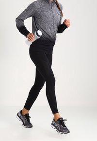 Zwarte sportleggings in combinatie met een grijze zip-up jacket en zwarte hardloopschoenen. Waterfles in één hand vastgehouden. Zachte stofstructuur.