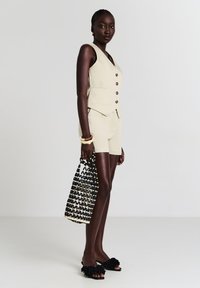 Frau in beiger ärmelloser Knopfveste und passender Shorts, die eine schwarz-weiße geflochtene Tasche hält, mit schwarzen floralen Sandalen und Armbändern.