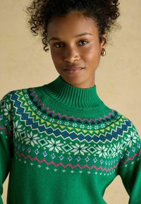 Grüner gestrickter Pullover mit hohem geripptem Kragen, der mehrfarbige geometrische Muster und weiße Schneeflocken entlang der Passe aufweist.