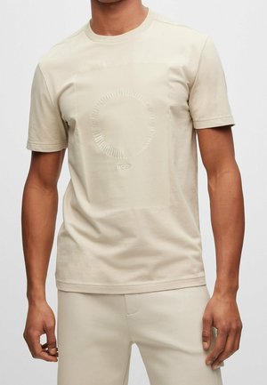 T-shirt beige clair en tissu doux, avec un logo circulaire et le texte "BOSS" au centre. Col rond classique, manches courtes.