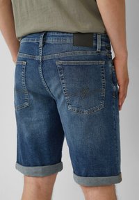 Shorts en denim dans un bleu moyen, avec un ourlet roulé, deux poches arrière avec des détails de coutures, et une étiquette de taille noire unie.