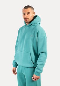 Sweat-shirt turquoise avec capuche et poche avant. Fabriqué en tissu doux, avec des poignets côtelés et un petit logo sur la poitrine.