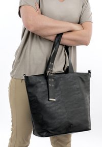 Asmuo sukryžiavęs rankas laiko didelę juodos odos rankinę su sagties detalė priešais smėlio spalvos kelnes ir šviesiai pilką marškinių foną.