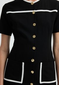 Czarna sukienka z krótkimi rękawami, złotymi guzikami, białą lamówką wzdłuż dekoltu i kieszeni, o dopasowanym kroju z dwiema przednimi kieszeniami.