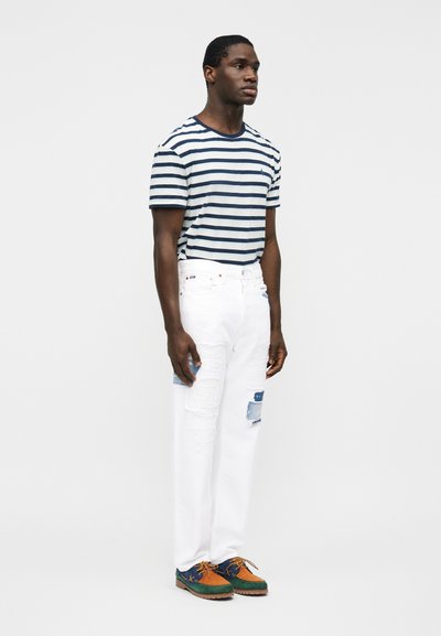 Άνδρας όρθιος πλαγίως, φορώντας μπλουζάκι με ναυτικό και λευκό ριγέ, λευκό τζιν με μπαλώματα και πολύχρωμα καφέ, πράσινα και ναυτικά παπούτσια.