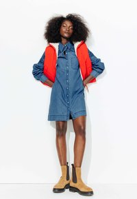 Robe chemise en denim avec un devant à boutons et un col, superposée avec un gilet rembourré rouge. Bottes cheville beiges avec des accents élastiques noirs et une semelle robuste.