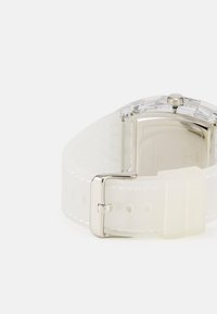 Montre en plastique transparent avec un boîtier rectangulaire en argent, bracelet en silicone blanc avec des découpes carrées, et fermeture à boucle en métal.