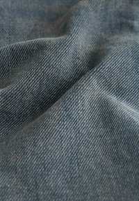 Denimstoff in einem mittleren Blauton, mit leichter Textur und sichtbarem Webmuster, das weiche Drapierungen und einen leichten Glanz zeigt.