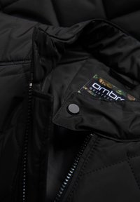 Sort quiltet jakke med en glat tekstur, der har en lynlåslukning fortil, trykknaplukning og et mærke synligt inden i kraven.