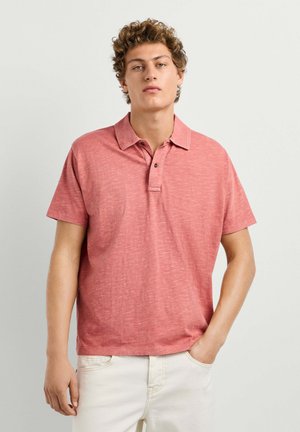 Junger Mann mit lockigem Haar, der ein kurzärmeliges pinkes Poloshirt und cremefarbene Hosen trägt, steht mit einer Hand in der Tasche.