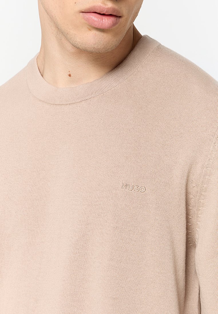 Beige gebreide sweater met een ronde hals en ribbelafwerking, met subtiele "HUGO" branding op de borst en textuurdetails op de mouwen.