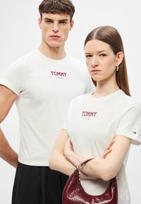 Λευκά βαμβακερά t-shirts με το λογότυπο "TOMMY JEANS" σε μπέργκουντι χρώμα. Το μοντέλο κρατά μια τσάντα χειρός με ανάγλυφο μοτίβο κροκοδειλέ, επίσης σε μπέργκουντι, και οι δύο βρίσκονται μπροστά σε ουδέτερο φόντο.