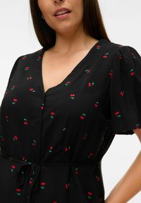 Robe noire avec un décolleté en V, des manches courtes bouffantes, ornée de petits motifs de cerises rouges et de feuilles vertes. Comprend une fermeture à boutons et une ceinture à nouer.