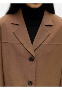 Bruine blazer met een gestructureerde kraag, drie zwarte knopen en een strak ontwerp. Draagt over een zwarte coltrui met een gladde textuur.