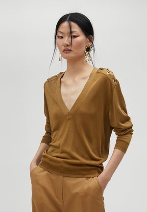 Brown V-neck sweater in soft fabric with textured accents and grommet details on the shoulders. Paired with tan high-waisted trousers.