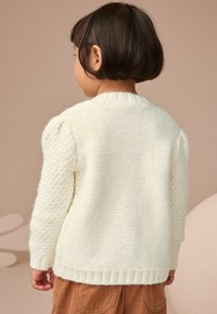 Cardigan en tricot couleur crème avec des manches texturées, un col et un ourlet côtelés, présentant une coupe décontractée et une apparence douce et confortable.