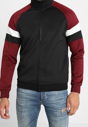 Mies, joka on pukeutunut mustavartiseen vetoketjulliseen takkiin, jossa on marjapuuron väriset hihat sekä valkoiset ja mustat raidat olkapäillä, yhdistettynä revittyihin sinisiin farkkuihin.