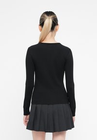 Vrouw met een blonde paardenstaart, gekleed in een zwart shirt met lange mouwen en een donkergrijze geplooide rok, staand met haar rug naar de camera tegen een witte achtergrond.