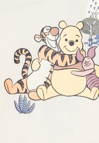 Witte stof met een afbeelding van drie cartoonfiguren: een lachende beer, een gestreepte tijger en een klein roze varken, met bladaccenten.