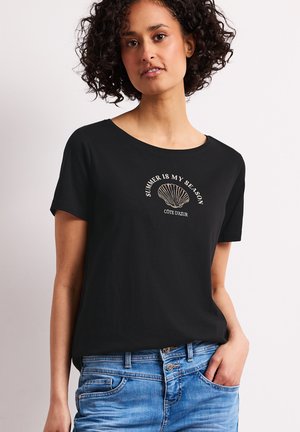 Femme aux cheveux bouclés et foncés portant un T-shirt noir avec un motif coquillage et le texte « Summer is my season Cote d'Azur », associé à un jean bleu.