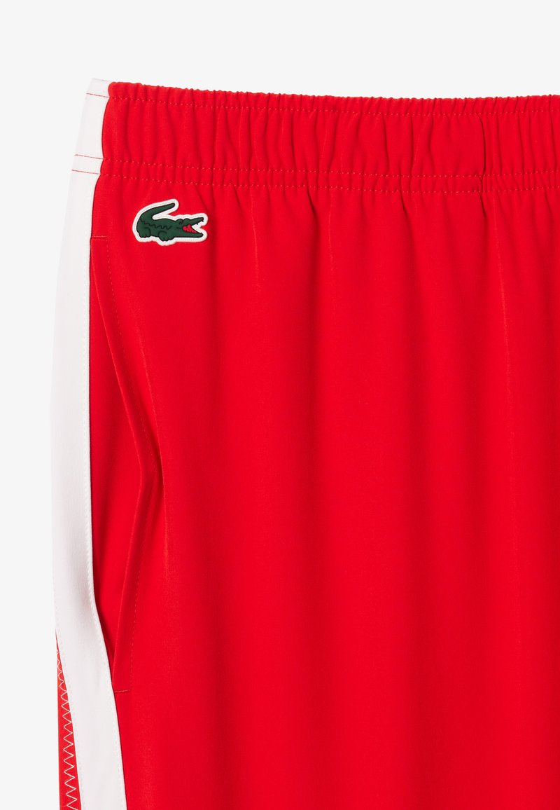 Shorts de sport rouges en tissu lisse, dotés d'une taille élastique et de bandes blanches sur les côtés ; comprend un logo Lacoste vert sur la hanche.