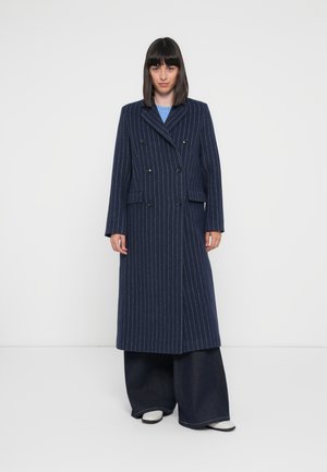 Long navy pinstripe coat with a double-breasted design, notched collar, and two front pockets. Paired with wide-leg denim pants and white shoes.
