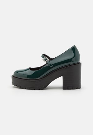 Green patent leather Mary Jane shoes with a thick black platform heel, rounded toe, and a buckle strap at the ankle. Textured sole.