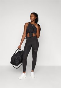 Ensemble de sport noir comprenant un haut court avec des volants et un legging taille haute. Le modèle tient un sac de sport noir et porte des baskets blanches.