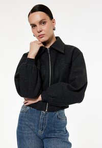 Sort bomberjakke i uldblanding med lynlås foran, stor krave og puffede ærmer; båret med lyseblå højtaljede jeans.