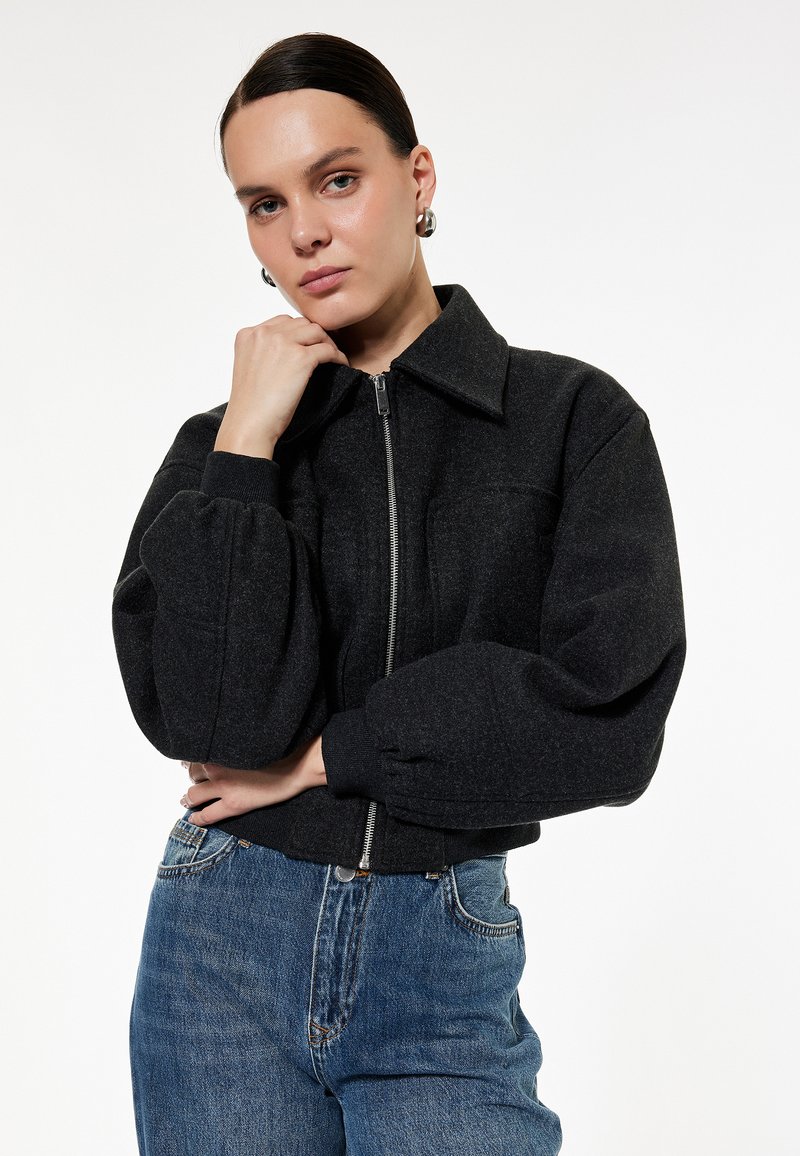 Sort bomberjakke i uldblanding med lynlås foran, stor krave og puffede ærmer; båret med lyseblå højtaljede jeans.