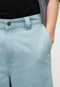 Person iført lyseblå denimshorts med knap og hånden delvist i højre lomme mod en ensfarvet baggrund.