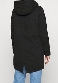 Svart isolerad jacka med huva, har en quiltad struktur, långa ärmar och en sidoklyfta i fållen. Bakifrån visar den en enkel design.