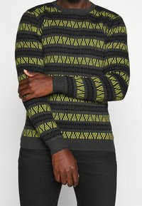 Strikket sweater med sorte og neongule geometriske mønstre, rund halsudskæring, ribstrikkede manchetter og en afslappet pasform. Materialet ser blødt og struktureret ud.