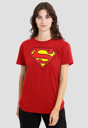 Woman wearing a red t-shirt with a large yellow and red Superman logo, standing against a plain light gray background.