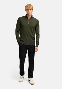 Olijfgroene pullover met een halve rits kraag, lange mouwen en geribbelde zoom, gecombineerd met zwarte broeken en witte sneakers met tan accenten.
