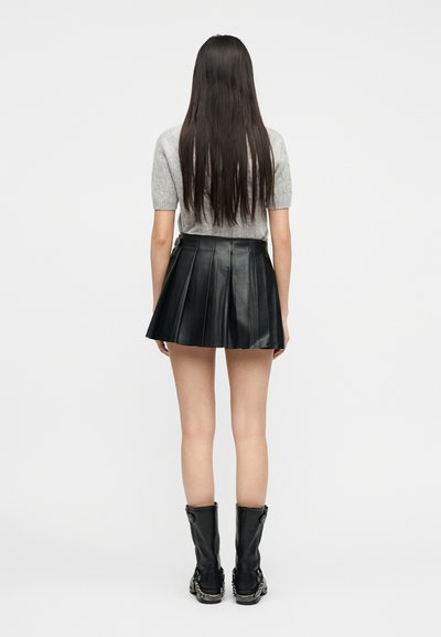 Femme aux longs cheveux bruns portant un haut gris à manches courtes, une mini-jupe plissée en cuir noir et des bottes noires mi-mollet avec des détails en chaîne, vue de dos.