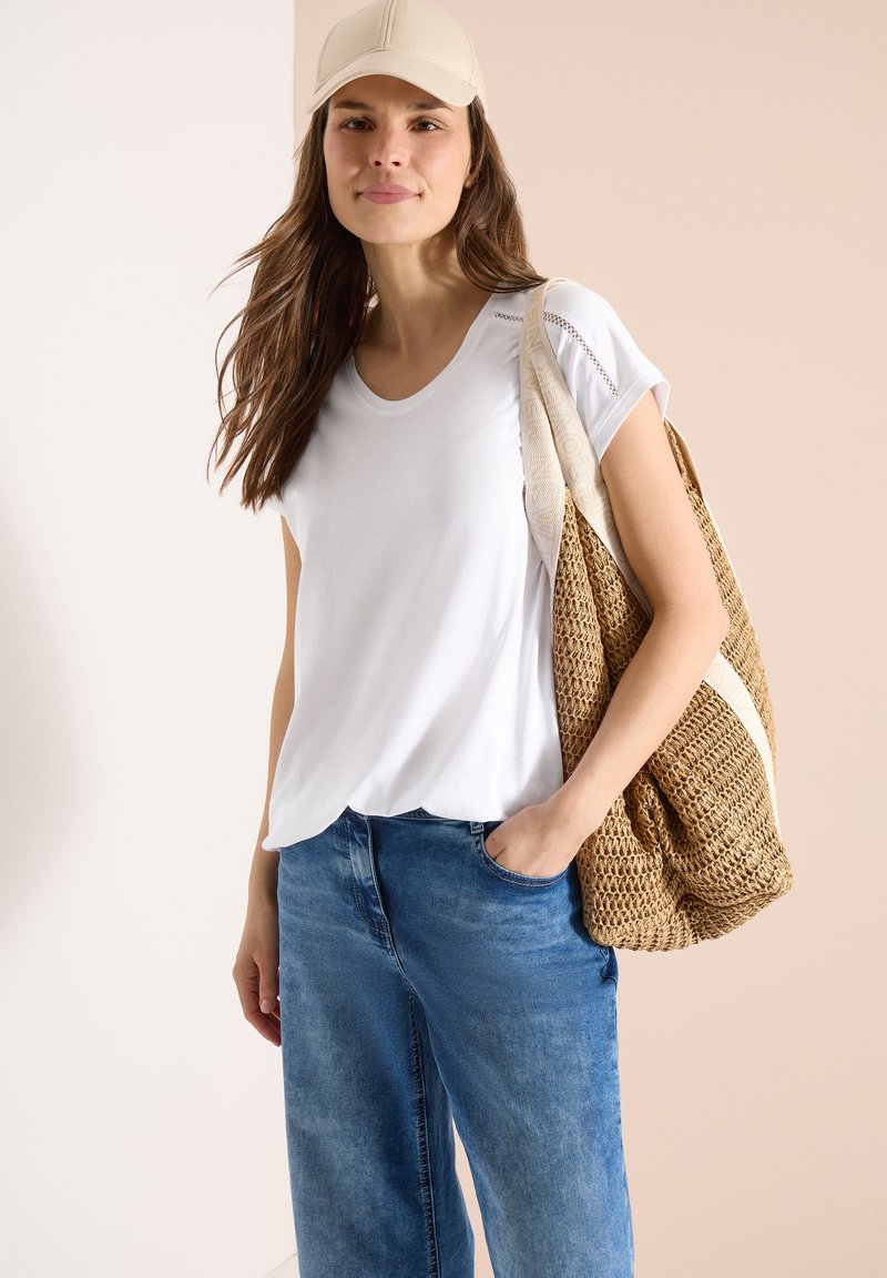 Femme portant une casquette beige, une chemise blanche à manches courtes, un jean bleu, tenant un grand sac fourre-tout tressé sur son épaule avec une main dans la poche.