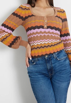 Femme portant un top en maille ajusté à manches longues avec des rayures en zigzag multicolores et trois petits boutons, assorti à un jean en denim bleu.
