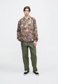 Barna terepszínű dzseki, alatta világoslila grafikás póló, olíva zöld nadrág és fekete sportcipő fehér fűzővel.