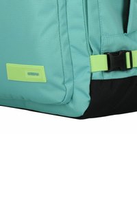 Ljus teal ryggsäck med svart botten. Har en gul-grön spänne och varumärkesetikett. Tillverkad av strukturerad textil med söm i detaljer.