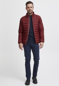 Giacca rossa trapuntata con colletto alto, chiusura con zip, tasche laterali; abbinata a pantaloni navy e scarpe nere. Materiale testurizzato e isolato.