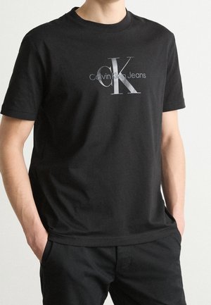 Osoba ubrana w czarny t-shirt Calvin Klein Jeans z szarym logo "ck" i napisem, ręce częściowo w kieszeniach, na jednolitym jasnym tle.