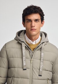 Giacca puffer trapuntata beige con cappuccio, con il marchio "POLO CLUB" sui lacci. Strato interno visibile in giallo, con una camicia colletto sotto.