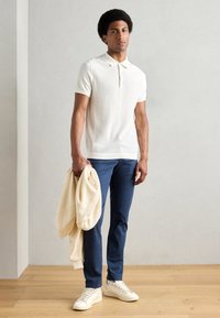 Polo shirt blanc à manches courtes et côtelé, chinos bleus, pull crème drapé sur le bras, et baskets blanches, debout sur un sol en bois.