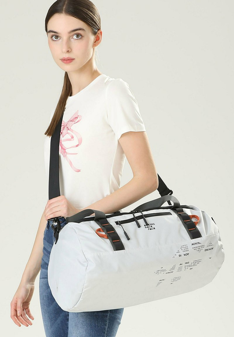 Sac de sport gris clair en matériau durable, avec des sangles noires, des accents orange, des fermetures éclair et un texte imprimé sur le côté.