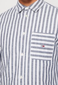 Koszula na guziki w niebiesko-białe paski. Posiada małą kieszonkę na piersi z logo marki. Materiał to teksturowana, lekka bawełna.