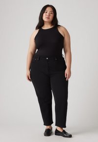 Débardeur noir à texture côtelée, associé à un jean taille haute noir avec poches avant, et des mocassins noirs sur un fond clair.